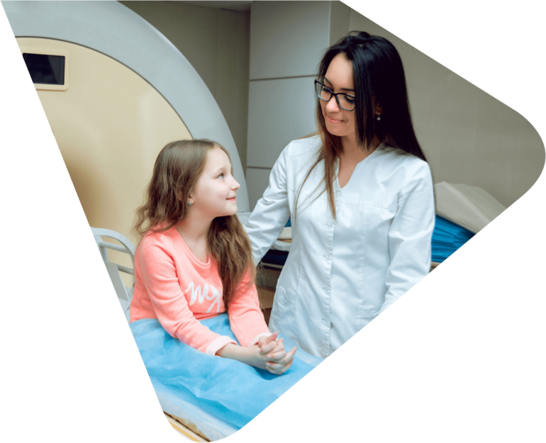 Young girl sitting on a medical scanning machine ready to be scanned, smiling up at a medical professional - a lady wearing a white lab coat with dark hair and glasses. Representing Woods Valldata's work with medical and health sector charities.