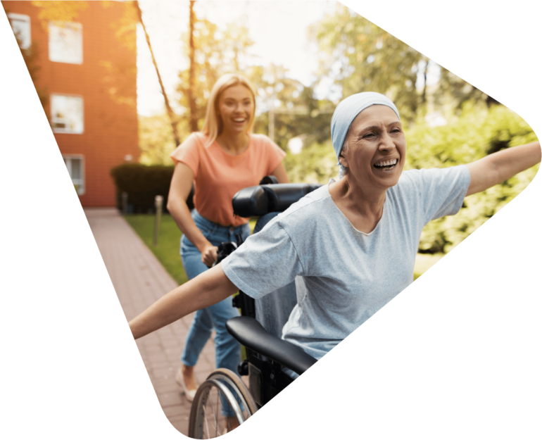 Cancer patient wearing a light blue headscarf and matching t-shirt sitting in a wheelchair with arms outstretched to the sides (like an aeroplane) smiling with joy and being pushed down a path by a young woman in blue jeans and orange t-shirt, with blond hair also smiling. Representing Woods Valldata's work with hospices and health sector charities.