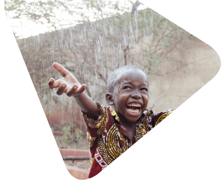 Young boy in traditional African print shirt smiling with his hands outstretched because it is raining. Representing Woods Valldata's work with international aid and disaster relief charities.