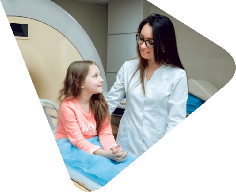 Young girl sitting on a medical scanning machine ready to be scanned, smiling up at a medical professional - a lady wearing a white lab coat with dark hair and glasses. Representing Woods Valldata's work with medical and health sector charities.