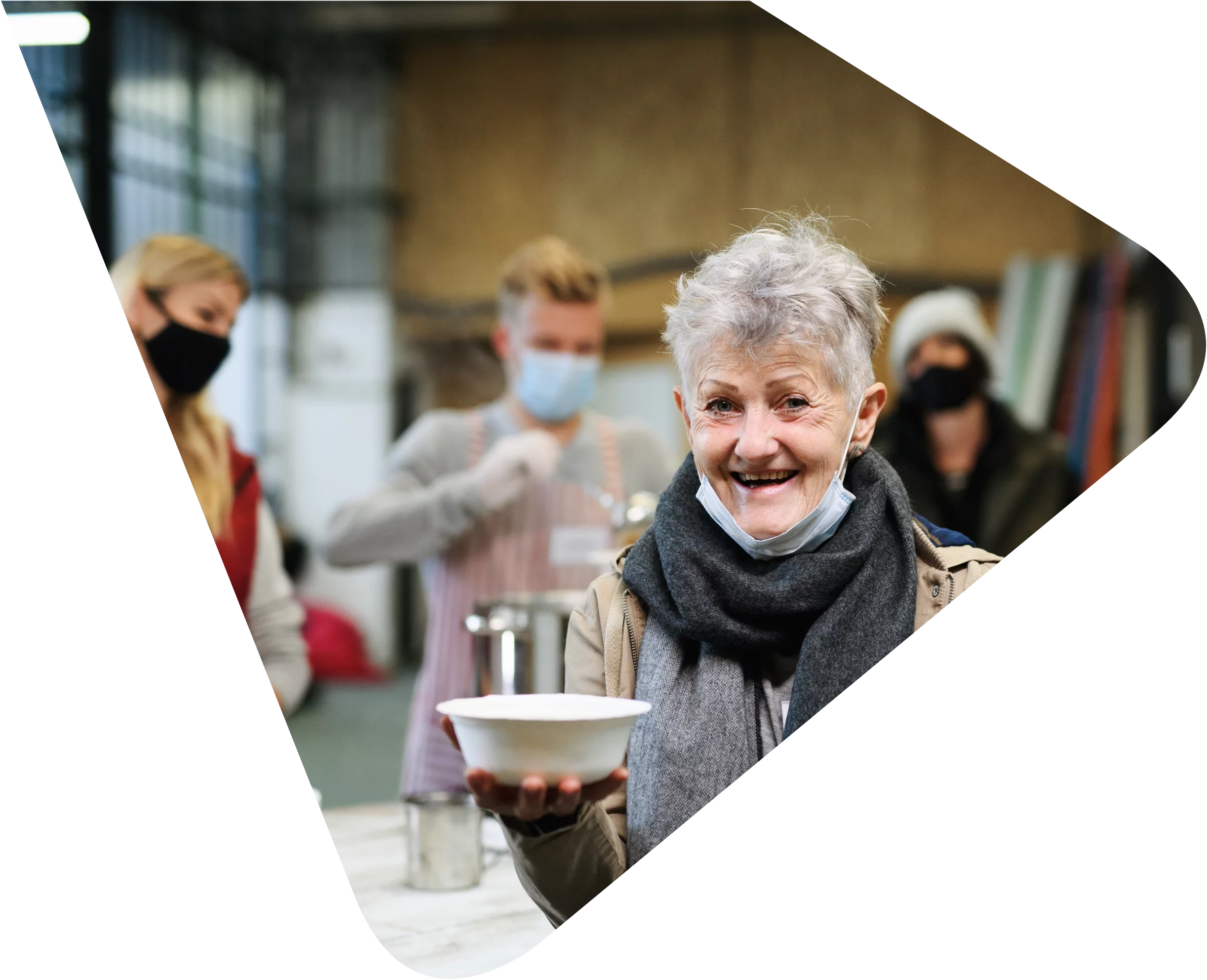 Image of an older homeless lady with short grey hair wearing a coat and scarf holding a white bowl. In the background are three servers in a soup kitchen wearing face masks. Representing Woods Valldata's work with community, homelessness and care sector charities.