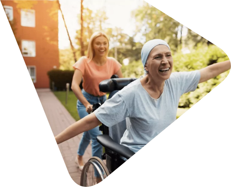 Cancer patient wearing a light blue headscarf and matching t-shirt sitting in a wheelchair with arms outstretched to the sides (like an aeroplane) smiling with joy and being pushed down a path by a young woman in blue jeans and orange t-shirt, with blond hair also smiling. Representing Woods Valldata's work with hospices and health sector charities.