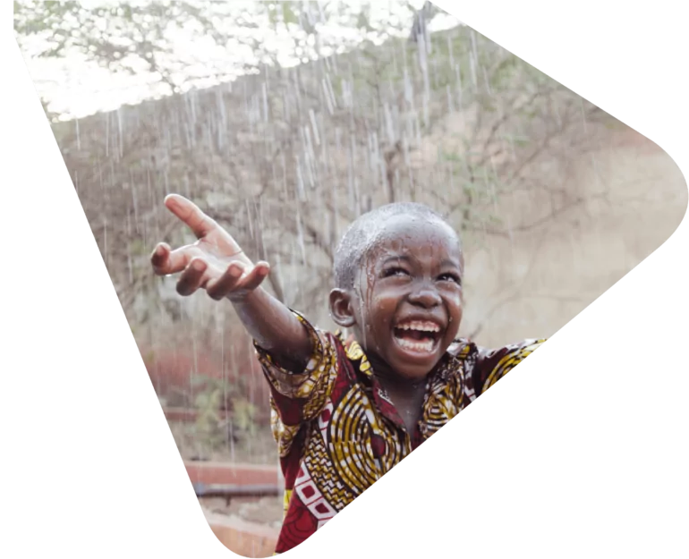 Young boy in traditional African print shirt smiling with his hands outstretched because it is raining. Representing Woods Valldata's work with international aid and disaster relief charities.